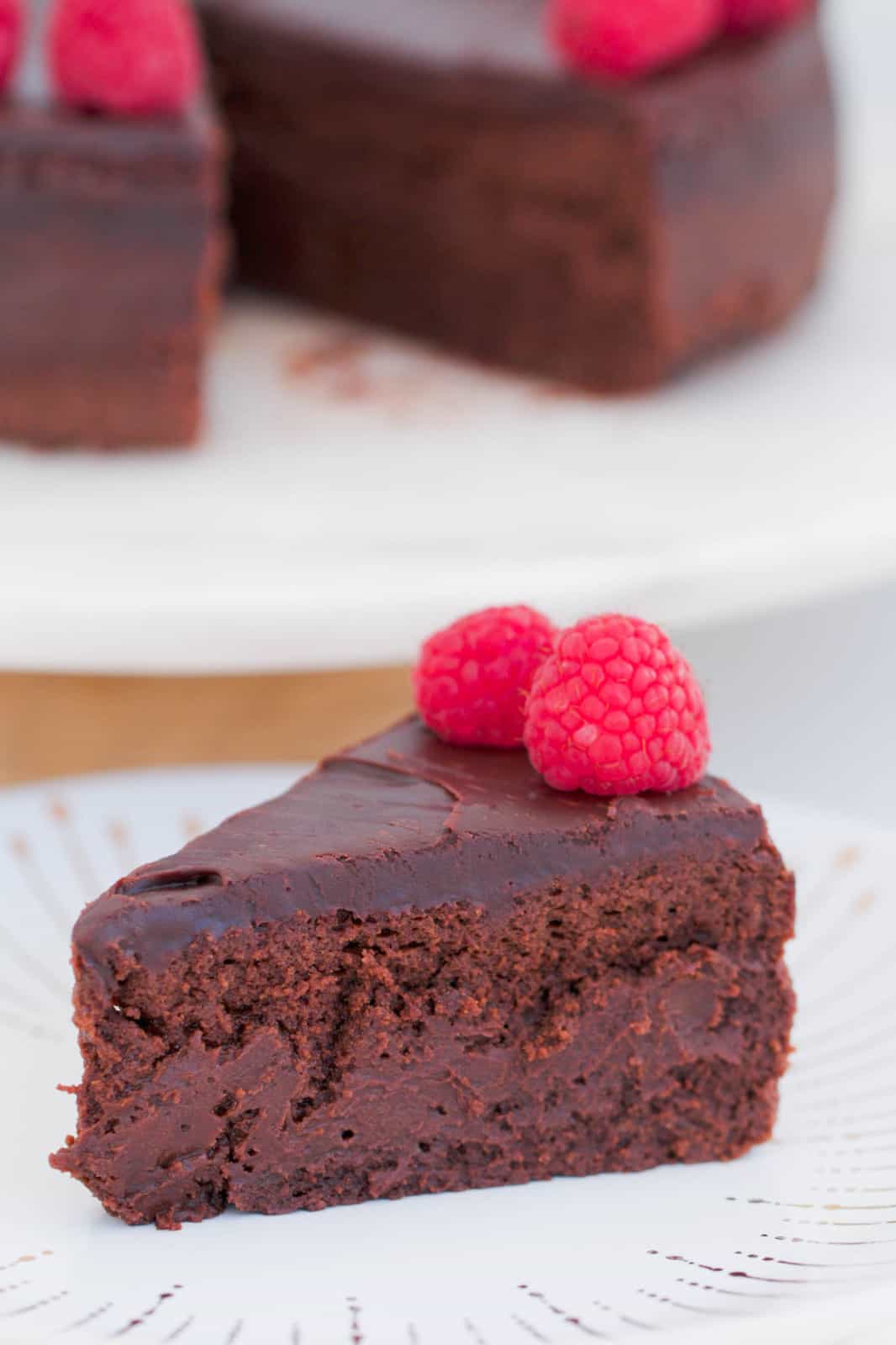 A slice of chocolate mousse cake topped with ganache and raspberries on a white plate.