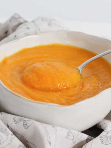 Carrot, Pumpkin & Sweet Potato Soup in a white bowl surrounded by a patterned tea towel